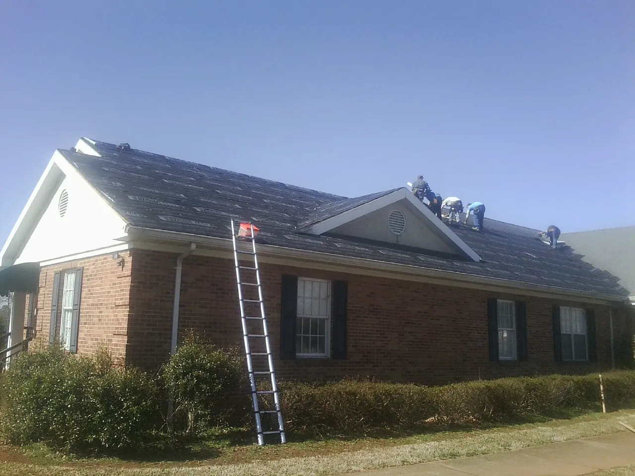 Crew tearing shingles off a brick ranch roof with a ladder set — Metal Roof Repair work by our crew in Helendale